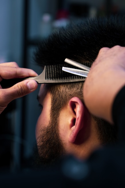 A close-up view of a person having a haircut, with a comb and scissors being used near the side of their head. The person has a well-groomed beard and short, dark hair. The focus is on the hands of the barber and the tools being used.