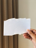 A hand is holding a tear-edged piece of white paper with a textured border. The background features a vertical brown curtain.