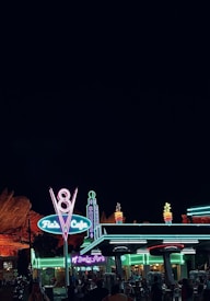 A diner with retro neon signs is illuminated against a dark sky. The signs feature bright colors and stylized fonts, with the surrounding area bustling with people. The architecture evokes a vintage feel with vibrant lights creating a glowing atmosphere.