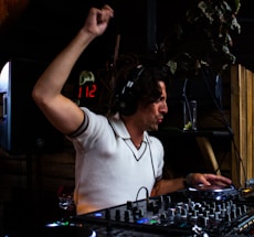 A DJ is energetically playing at a set, wearing headphones and a white shirt. The DJ appears focused and expressive, with equipment such as a mixer and turntable in the foreground. Dim lighting and a wooden interior provide a cozy atmosphere.
