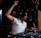 A DJ is energetically playing at a set, wearing headphones and a white shirt. The DJ appears focused and expressive, with equipment such as a mixer and turntable in the foreground. Dim lighting and a wooden interior provide a cozy atmosphere.