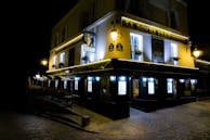 A two-story building at night is illuminated by multiple lights, including wall-mounted lamps and bright signs. The building has an inviting atmosphere with flower pots under the windows and glowing windows indicating activity inside. The signage prominently displays 'La Bonne Franquette'. The surrounding area features cobblestone streets, giving it a classic, old-world charm.