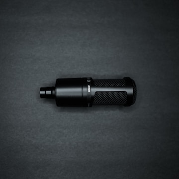 A black cylindrical microphone with a mesh pattern on its body sits against a dark background. The microphone has a sleek, modern design with a matte finish and visible text or branding on the side.
