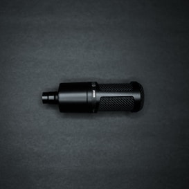 A black cylindrical microphone with a mesh pattern on its body sits against a dark background. The microphone has a sleek, modern design with a matte finish and visible text or branding on the side.