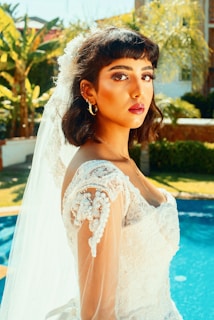 A young woman is wearing a bridal gown with intricate lace details and beading. Her dark hair is styled in loose waves, and she has a long veil extending down her back. The setting appears to be outdoors by a pool, with lush greenery in the background.
