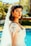 A young woman is wearing a bridal gown with intricate lace details and beading. Her dark hair is styled in loose waves, and she has a long veil extending down her back. The setting appears to be outdoors by a pool, with lush greenery in the background.