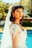 A young woman is wearing a bridal gown with intricate lace details and beading. Her dark hair is styled in loose waves, and she has a long veil extending down her back. The setting appears to be outdoors by a pool, with lush greenery in the background.