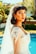 A young woman is wearing a bridal gown with intricate lace details and beading. Her dark hair is styled in loose waves, and she has a long veil extending down her back. The setting appears to be outdoors by a pool, with lush greenery in the background.
