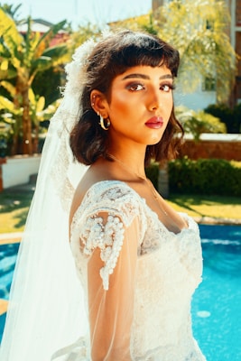 A young woman is wearing a bridal gown with intricate lace details and beading. Her dark hair is styled in loose waves, and she has a long veil extending down her back. The setting appears to be outdoors by a pool, with lush greenery in the background.