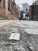 A small salt packet lies on cobblestone pavement in an urban alleyway. The background features blurred buildings with brick exteriors, characteristic of a city setting. The perspective is low to the ground, emphasizing the texture of the cobblestones.