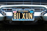 A vintage California license plate with the number 811 XUN is mounted on a classic car. The frame around the license plate is metallic, and the plate displays registration information, including 'Vallejo' and 'Earl A. Jensen'. The chrome bumper and grille of the car are visible, reflecting light and emphasizing the antique style.