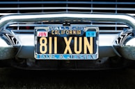 A vintage California license plate with the number 811 XUN is mounted on a classic car. The frame around the license plate is metallic, and the plate displays registration information, including 'Vallejo' and 'Earl A. Jensen'. The chrome bumper and grille of the car are visible, reflecting light and emphasizing the antique style.