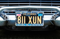 A vintage California license plate with the number 811 XUN is mounted on a classic car. The frame around the license plate is metallic, and the plate displays registration information, including 'Vallejo' and 'Earl A. Jensen'. The chrome bumper and grille of the car are visible, reflecting light and emphasizing the antique style.