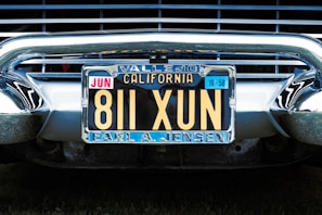 A vintage California license plate with the number 811 XUN is mounted on a classic car. The frame around the license plate is metallic, and the plate displays registration information, including 'Vallejo' and 'Earl A. Jensen'. The chrome bumper and grille of the car are visible, reflecting light and emphasizing the antique style.