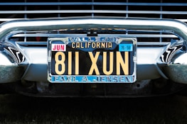 A vintage California license plate with the number 811 XUN is mounted on a classic car. The frame around the license plate is metallic, and the plate displays registration information, including 'Vallejo' and 'Earl A. Jensen'. The chrome bumper and grille of the car are visible, reflecting light and emphasizing the antique style.