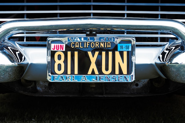 A vintage California license plate with the number 811 XUN is mounted on a classic car. The frame around the license plate is metallic, and the plate displays registration information, including 'Vallejo' and 'Earl A. Jensen'. The chrome bumper and grille of the car are visible, reflecting light and emphasizing the antique style.