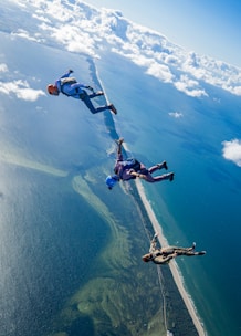 Three skydivers are freefalling above a coastal landscape with clear blue skies and scattered clouds. The ocean and shoreline are visible below, creating a breathtaking view from high altitude.