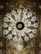 An ornate chandelier with intricate glass designs hangs from an elaborately decorated ceiling. The chandelier features leafy glass elements and blue accents, with a symmetrical pattern radiating from its center. The ceiling itself is adorned with gilded molding and a classical painting, adding to the luxurious atmosphere.