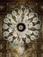 An ornate chandelier with intricate glass designs hangs from an elaborately decorated ceiling. The chandelier features leafy glass elements and blue accents, with a symmetrical pattern radiating from its center. The ceiling itself is adorned with gilded molding and a classical painting, adding to the luxurious atmosphere.