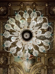 An ornate chandelier with intricate glass designs hangs from an elaborately decorated ceiling. The chandelier features leafy glass elements and blue accents, with a symmetrical pattern radiating from its center. The ceiling itself is adorned with gilded molding and a classical painting, adding to the luxurious atmosphere.