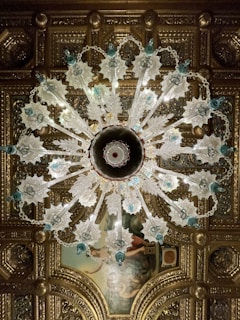 An ornate chandelier with intricate glass designs hangs from an elaborately decorated ceiling. The chandelier features leafy glass elements and blue accents, with a symmetrical pattern radiating from its center. The ceiling itself is adorned with gilded molding and a classical painting, adding to the luxurious atmosphere.