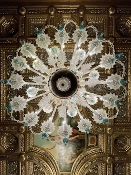 An ornate chandelier with intricate glass designs hangs from an elaborately decorated ceiling. The chandelier features leafy glass elements and blue accents, with a symmetrical pattern radiating from its center. The ceiling itself is adorned with gilded molding and a classical painting, adding to the luxurious atmosphere.
