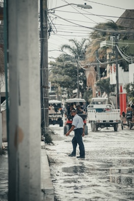 A man walks through a flooded street lined with power lines and palm trees. Several vehicles and people are visible in the background, navigating the wet and reflective surface of the road. The setting appears urban, with buildings flanking the street.