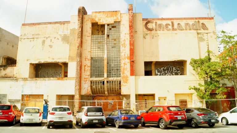 An old, abandoned cinema building with a weathered facade and a faded sign that reads 'CineLandia'. The structure is in a state of disrepair, with rust and peeling paint visible. Several cars are parked in front of the building in a lined parking lot. Graffiti is present on the windows, adding to the feeling of neglect.