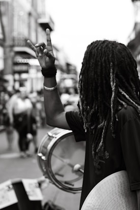 A person with long dreadlocks, wearing a dark shirt, holds up their hand in a rock or peace gesture in a bustling street. They are holding drums or cymbals, suggesting they might be part of a street performance. The street is busy with people walking around, indicating a lively and possibly urban setting.