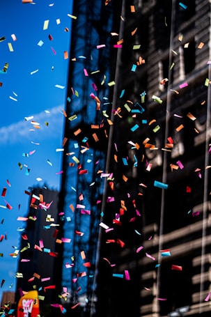 Multicolored confetti is suspended in mid-air against a backdrop of a tall building and a clear blue sky. The scene is vibrant and dynamic, with the confetti appearing to float and tumble freely.
