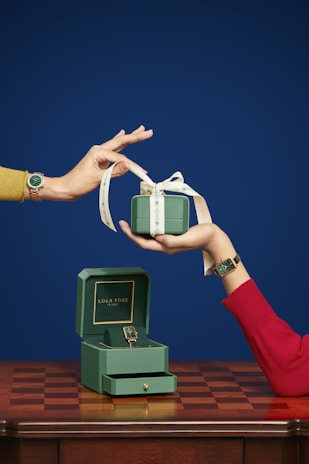 A green gift box with a ribbon is being handed over by a person wearing a red sleeve. The box is complemented by a matching green watch on the person's wrist. Below, there is an open gift box labeled 'Lola Rose,' containing another green watch, resting on a wooden table with a checkered pattern.