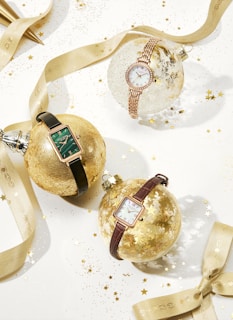 Three wristwatches are elegantly displayed on decorative Christmas ornaments. The watches vary in design with different strap colors and shapes. They are placed among a festive arrangement of ribbons and scattered gold stars, creating a luxurious and celebratory atmosphere.
