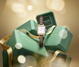 Several green hexagonal boxes are stacked with a brown leather wristwatch in front. The boxes bear the logo 'Lola Rose.' A gold ribbon is elegantly draped across them, enhancing a luxurious feel with soft, blurred golden lights in the background.