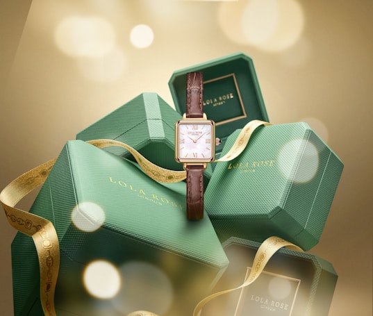 Several green hexagonal boxes are stacked with a brown leather wristwatch in front. The boxes bear the logo 'Lola Rose.' A gold ribbon is elegantly draped across them, enhancing a luxurious feel with soft, blurred golden lights in the background.