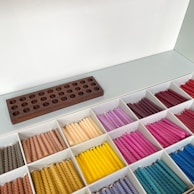 A neatly organized display of colorful twisted candles arranged in a grid of white compartments. The candles are grouped by color, including shades of brown, yellow, pink, purple, red, and blue. A dark wooden holder with multiple circular slots rests above the compartments.