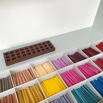 A neatly organized display of colorful twisted candles arranged in a grid of white compartments. The candles are grouped by color, including shades of brown, yellow, pink, purple, red, and blue. A dark wooden holder with multiple circular slots rests above the compartments.