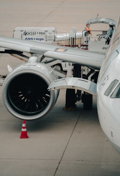 A close-up view of an airplane on a tarmac, focusing on the engine and wing. A cargo container marked ANA Cargo is in the background with two workers nearby. An orange safety cone is placed near the engine.