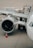 A close-up view of an airplane on a tarmac, focusing on the engine and wing. A cargo container marked ANA Cargo is in the background with two workers nearby. An orange safety cone is placed near the engine.