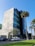 A modern multi-story office building with a facade featuring large glass windows and concrete panels. In the foreground, there are several palm trees and a few parked cars along a street. The sky is clear and blue, and the grass on the lawn is vibrant and well-maintained.
