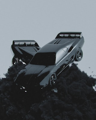A sleek, futuristic car seems to be floating or emerging from dark, billowing smoke against a muted gray background. The car has a shiny, reflective surface and aerodynamic design, emphasizing speed and modernity.