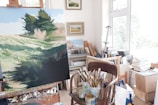 A bright, well-lit art studio filled with various paintings, paintbrushes, and art supplies. An easel holds a canvas painted with a landscape depicting trees and grassy fields. Several framed artworks and canvases are leaned against the walls. Natural light floods the room through large windows, and numerous art supplies and tools are scattered around.