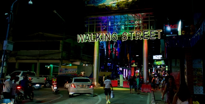 A bustling street scene at night with a large illuminated sign reading 'Walking Street'. The area is lively with people walking around, and vehicles including cars and motorcycles are visible on the road. Neon lights and advertisements create a vibrant and colorful atmosphere.