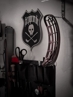 A wall-mounted tool organizer holds various tools including scissors and screwdrivers. Above it, a decorative plaque features a skull with two crossed hockey sticks and the phrase 'Fat pipe or die'. Additionally, a curved metallic object is attached to the wall.