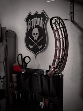 A wall-mounted tool organizer holds various tools including scissors and screwdrivers. Above it, a decorative plaque features a skull with two crossed hockey sticks and the phrase 'Fat pipe or die'. Additionally, a curved metallic object is attached to the wall.
