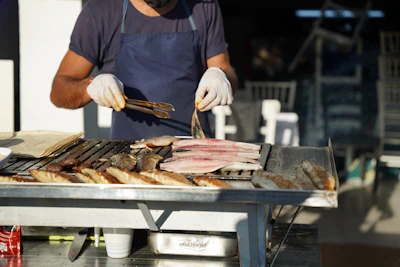A person wearing gloves handles fish fillets and whole fish cooking on an outdoor grill. The setup appears to be in a casual dining or market environment, with other items like a stack of flatbreads and disposable cups nearby.