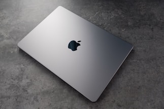 A sleek, silver laptop with a prominent logo on top is resting on a textured, grey surface. The laptop appears closed and is positioned at an angle that highlights its thin design.