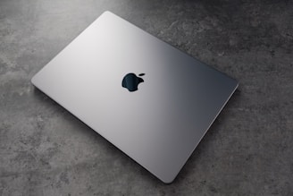A sleek, silver laptop with a prominent logo on top is resting on a textured, grey surface. The laptop appears closed and is positioned at an angle that highlights its thin design.