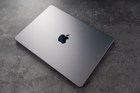 A sleek, silver laptop with a prominent logo on top is resting on a textured, grey surface. The laptop appears closed and is positioned at an angle that highlights its thin design.