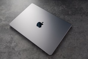A sleek, silver laptop with a prominent logo on top is resting on a textured, grey surface. The laptop appears closed and is positioned at an angle that highlights its thin design.