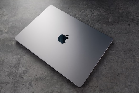 A sleek, silver laptop with a prominent logo on top is resting on a textured, grey surface. The laptop appears closed and is positioned at an angle that highlights its thin design.